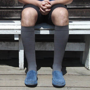 A person seated on a weathered wooden bench wearing dark gray knee-high socks and blue slip-on shoes, photographed outdoors on a wooden deck. - okotoks hc pharmacy