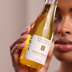 Bottle of Eminence Organic Skin Care Apricot Body Oil held by a hand with a blurred face in the background.