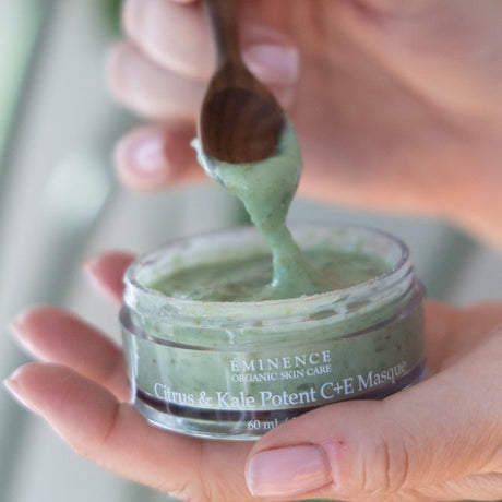 Hand holding a jar of Eminence Citrus & Kale Potent C+E Masque with a wooden spoon. - okotoks hc pharmacy