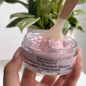 Hand holding a jar of Eminence Strawberry Khubarb Masque with a wooden applicator, plant in the background. - okotoks hc pharmacy