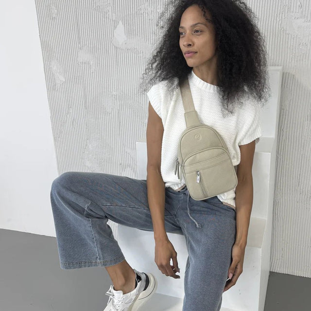 Woman sitting on a white chair wearing a white top, blue jeans, and a beige crossbody bag. - okotoks hc pharmacy