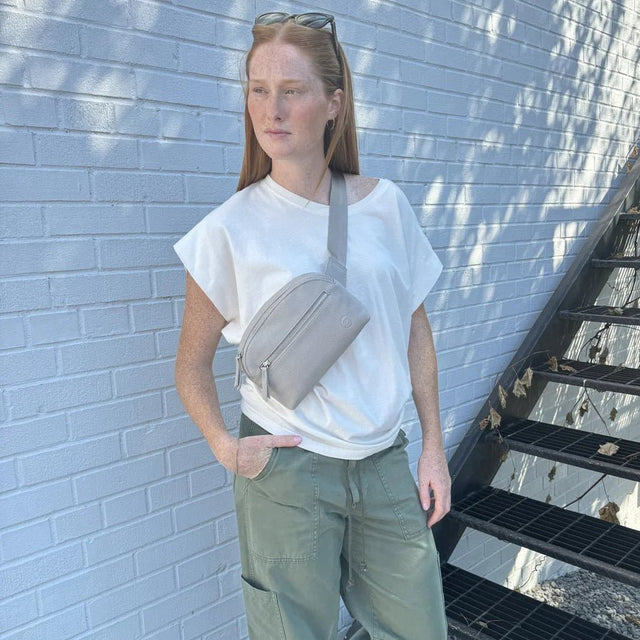 Person wearing a white t-shirt and green pants with a beige crossbody bag against a light blue brick wall. - okotoks hc pharmacy