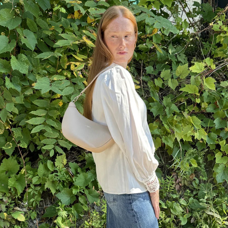 Woman holding a beige handbag in front of green foliage - okotoks hc pharmacy