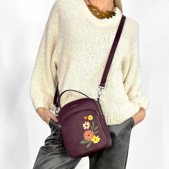 Person wearing a cream sweater and gray pants with a burgundy handbag featuring floral embroidery on a white background - okotoks hc pharmacy