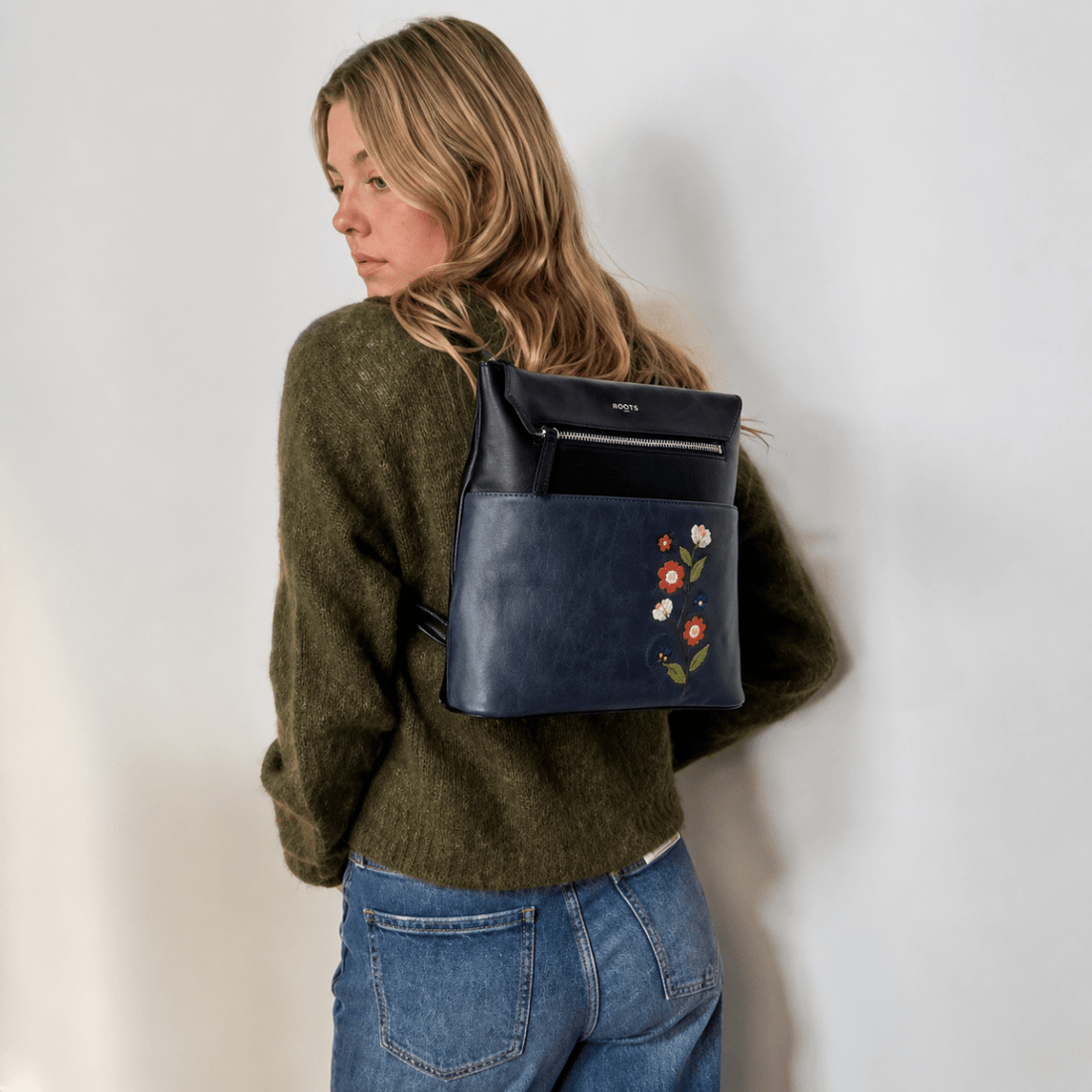 Person wearing a green sweater and blue jeans with a navy backpack featuring floral embroidery. - okotoks hc pharmacy