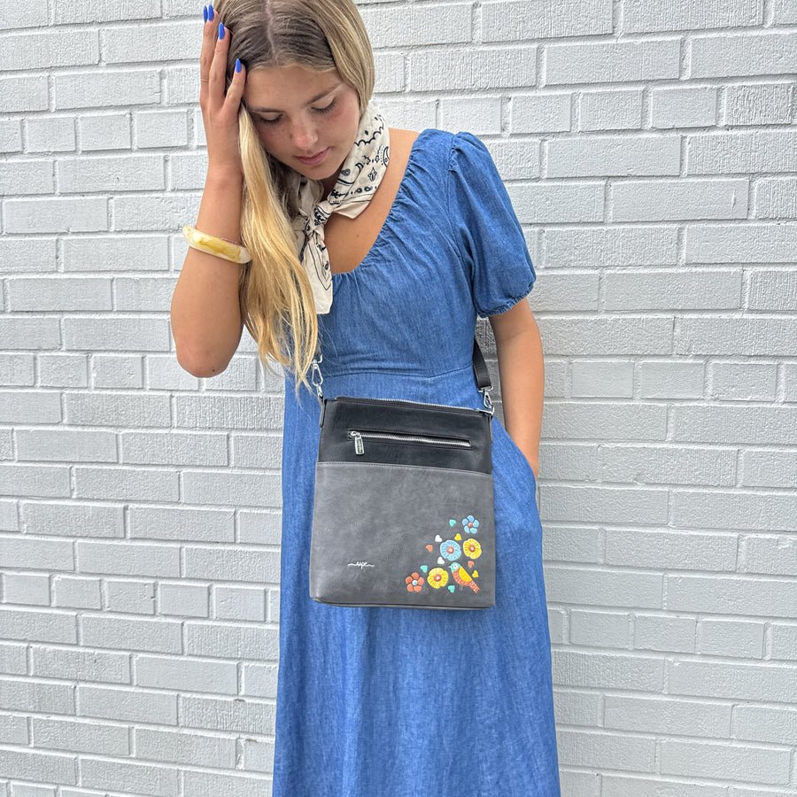 Woman in a blue dress holding a black handbag with floral designs against a gray brick wall. - okotoks hc pharmacy