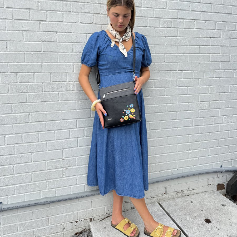 Woman in a blue dress holding a black handbag with colorful embellishments against a white brick wall. - okotoks hc pharmacy