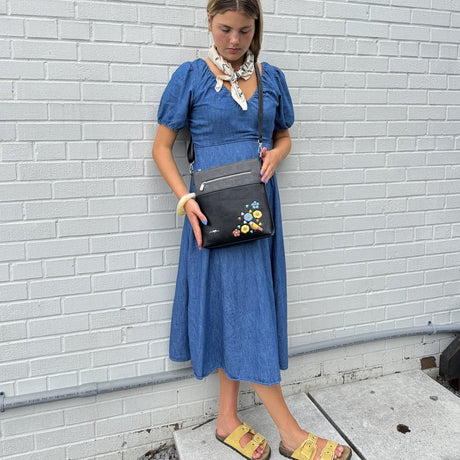 Woman in a blue dress holding a black handbag with colorful embellishments against a white brick wall. - okotoks hc pharmacy