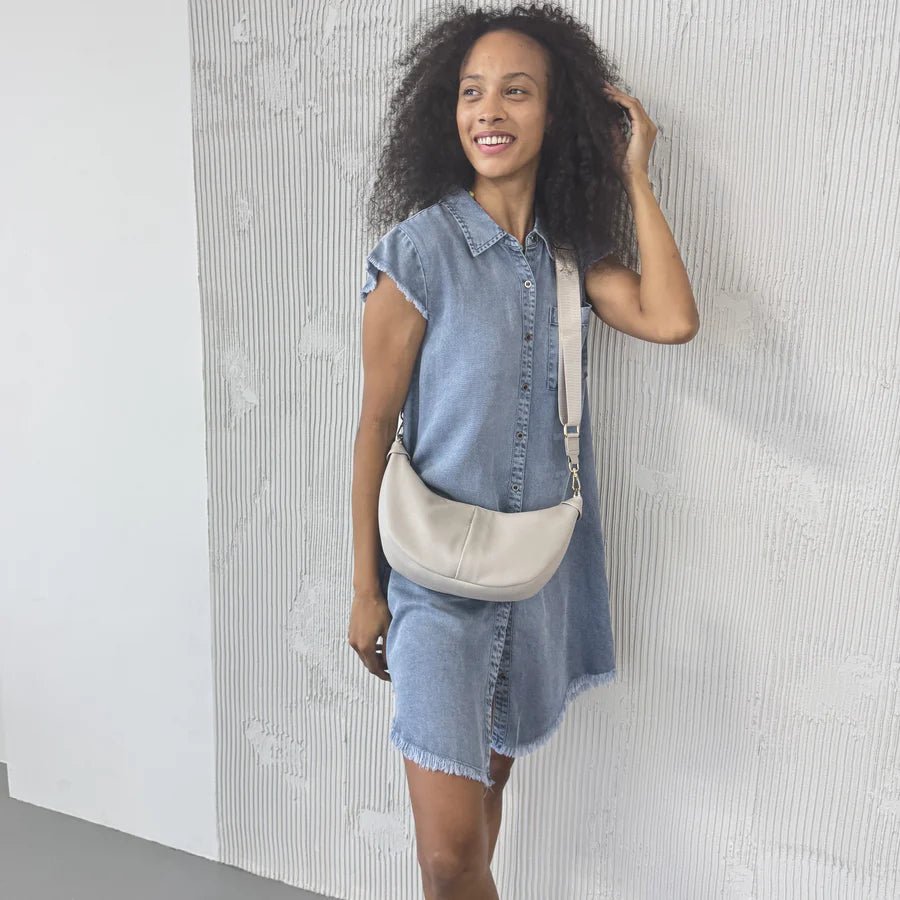 Woman wearing a denim dress with a beige bag against a textured white wall - okotoks hc pharmacy