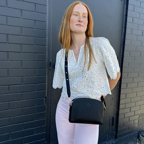 Woman wearing a white blouse and holding a black handbag against a brick wall. - okotoks hc pharmacy