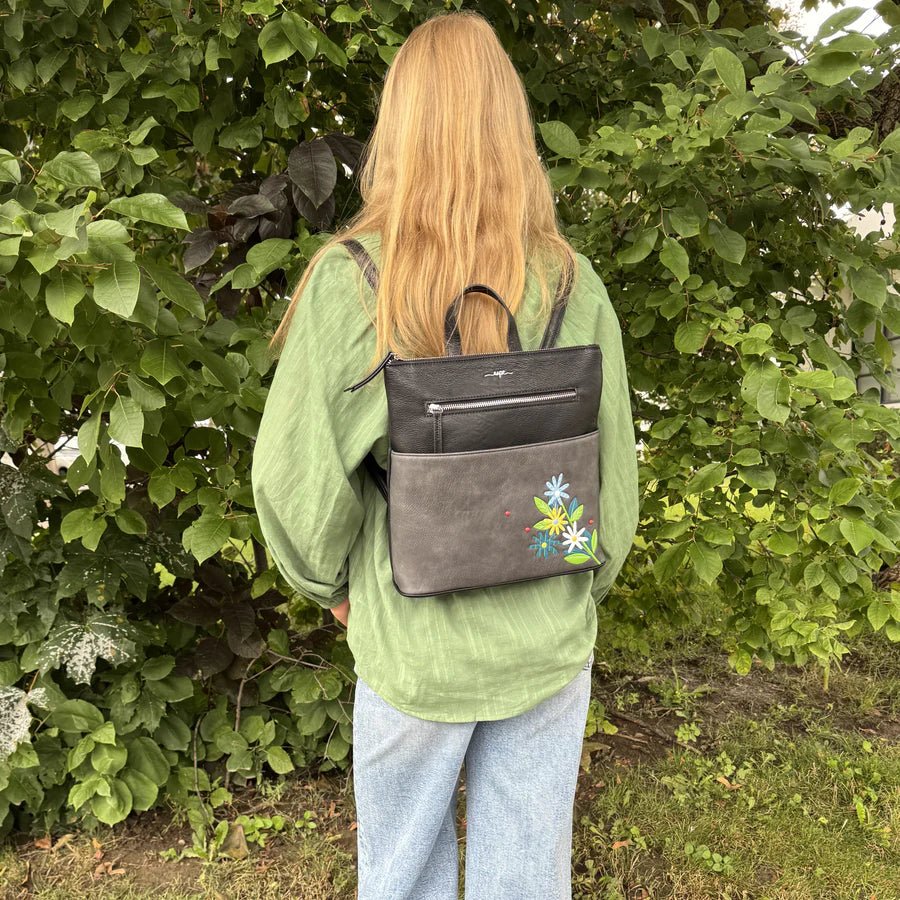Person wearing a green jacket with a gray backpack featuring floral designs, standing in front of green foliage. - okotoks hc pharmacy