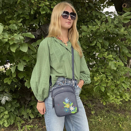 Woman wearing a green shirt and sunglasses, holding a gray crossbody bag with floral designs, standing in front of green foliage. - okotoks hc pharmacy