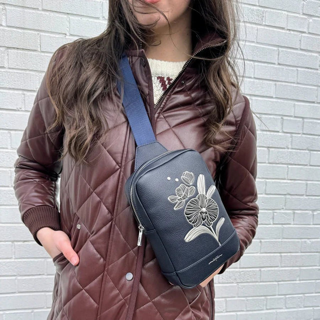 Person wearing a brown quilted jacket with a blue crossbody bag featuring floral design against a light gray brick wall. - okotoks hc pharmacy