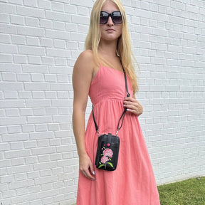 Woman in a pink dress holding a black crossbody bag with floral design against a white brick wall. - okotoks hc pharmacy