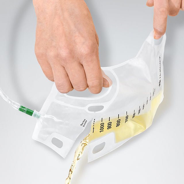 A clear plastic fluid collection bag with measurement markings and a green connector, being held open by two hands while a small amount of pale yellow liquid is poured into it, on a light gray background. - okotoks hc pharmacy