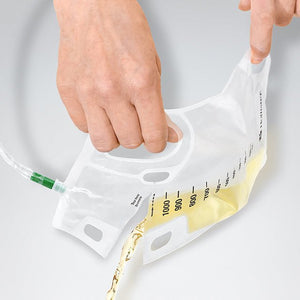 A clear plastic fluid collection bag with measurement markings and a green connector, being held open by two hands while a small amount of pale yellow liquid is poured into it, on a light gray background. - okotoks hc pharmacy