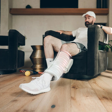 A man sits in a dark armchair in a living room while wearing a white and gray electronic leg stimulator device on his lower leg, with a white Nike Air Max sneaker visible. - okotoks hc pharmacy