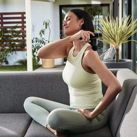Woman using a handheld massager on her arm while sitting on a couch outdoors. - okotoks hc pharmacy