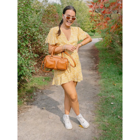 A woman stands on a dirt path outdoors wearing a yellow floral mini dress, a tan leather handbag, white sneakers, and sunglasses, with green foliage and trees on both sides of the path. - okotoks hc pharmacy