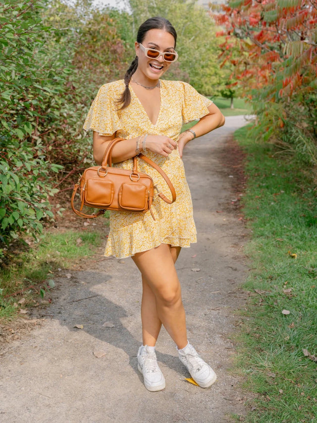 A woman stands on a dirt path outdoors wearing a yellow floral mini dress, a tan leather handbag, white sneakers, and sunglasses, with green foliage and trees on both sides of the path. - okotoks hc pharmacy
