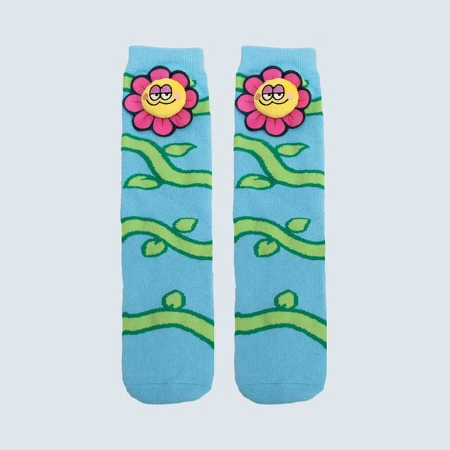 A pair of light blue crew socks featuring a cartoon floral pattern with pink flowers, yellow smiling faces, and green vines on a light gray background. - okotoks hc pharmacy