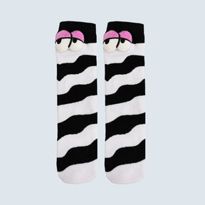 A pair of novelty socks featuring a black and white wavy zebra-like stripe pattern, with a cartoon face detail at the top showing pink eyebrows and white eyes, set against a solid light gray background. - okotoks hc pharmacy