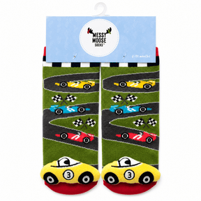 A pair of children's socks with a racing theme, hanging from a light blue cardboard hanger. The socks feature a green grass-like background with winding gray tracks, colorful race cars (yellow, blue, red), and checkered flags, with yellow cars at the toes on a red footbed. - okotoks hc pharmacy