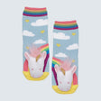 A pair of children's socks featuring a colorful unicorn design with a white face, pink mane, yellow horn, and rainbow tail on a light blue background with clouds and stars. The socks have rainbow-striped cuffs and yellow toes, shown against a light gray background. - okotoks hc pharmacy