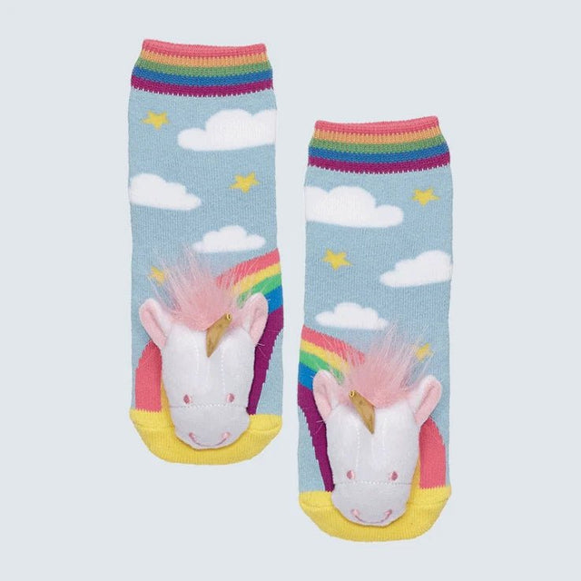 A pair of children's socks featuring a colorful unicorn design with a white face, pink mane, yellow horn, and rainbow tail on a light blue background with clouds and stars. The socks have rainbow-striped cuffs and yellow toes, shown against a light gray background. - okotoks hc pharmacy