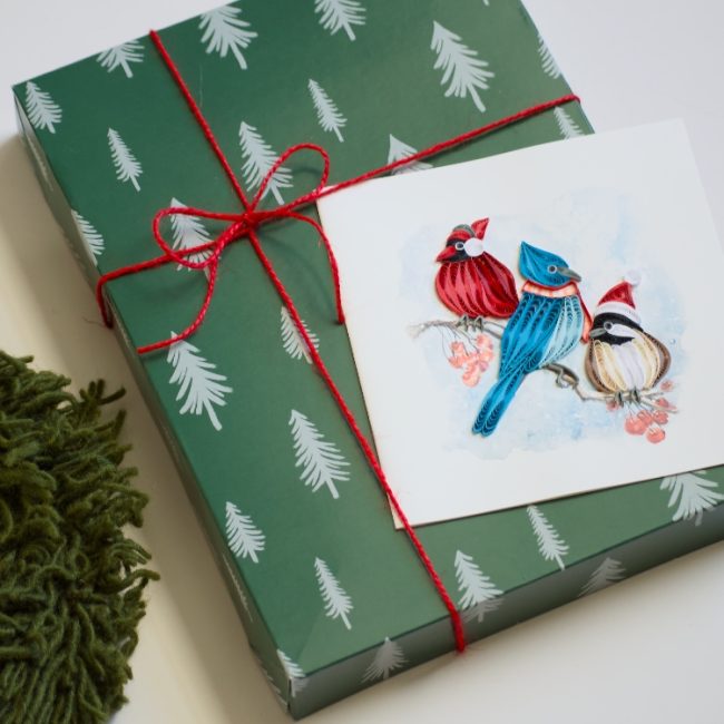Green gift box with red string, Christmas card with birds, and greenery on a white background - okotoks hc pharmacy